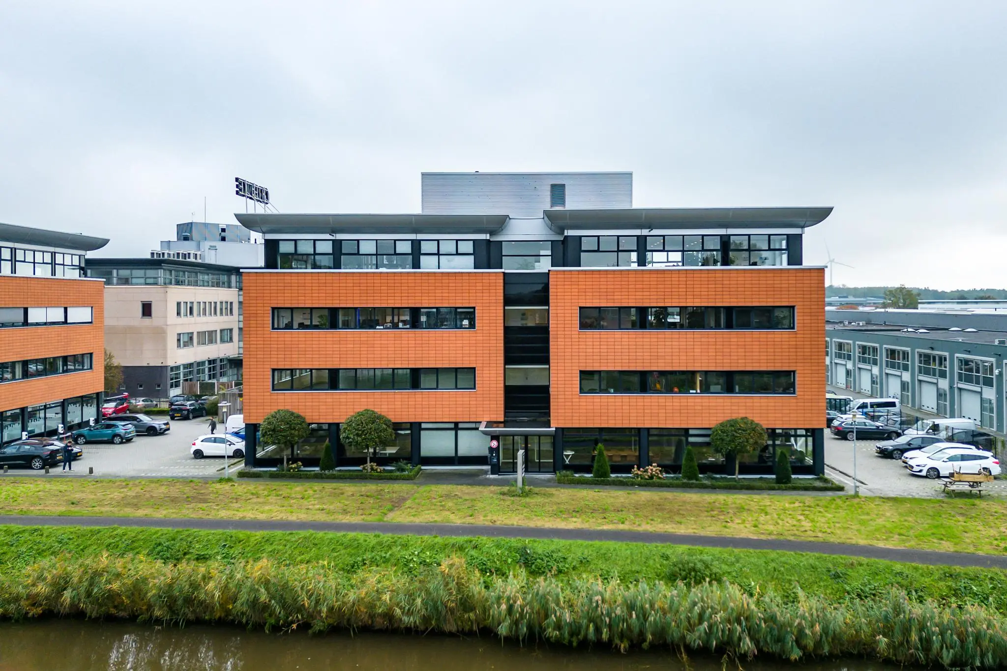Modern kantoorpand aan de Lage Biezenweg met oranje gevelbekleding en omliggende parkeerplaatsen, gezien vanaf de overkant van het water.